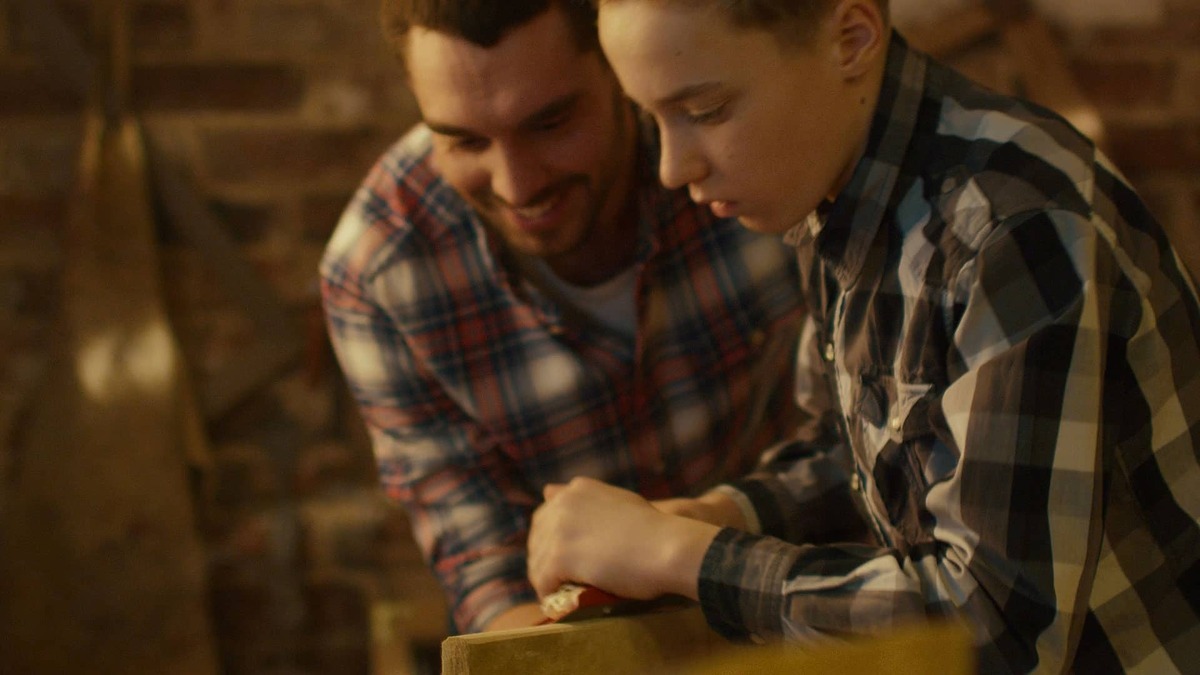 Mann und Kind bearbeiten in Werkstatt ein Stück Holz mit Hobel